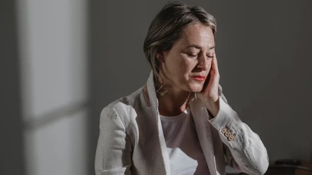 Woman in her early 50s with eyes closed and hand pressed to her cheek in quiet distress, absorbing the realization that her nightly melatonin dose may still be active in her bloodstream hours after waking.