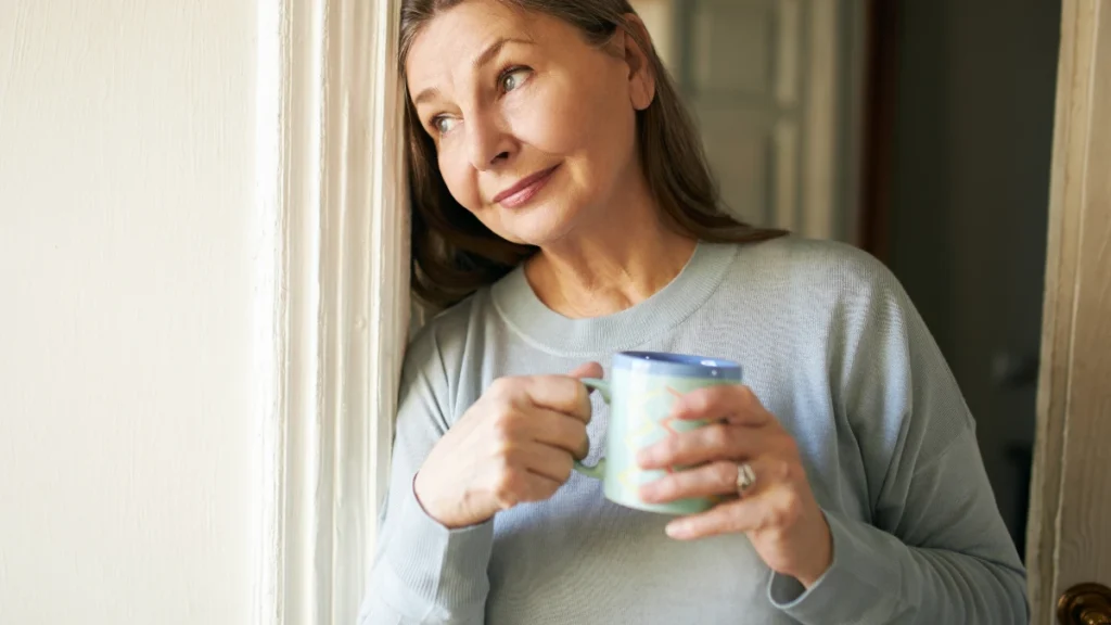 Woman in her late 50s leaning against a door frame holding a painted ceramic mug, gazing upward with a calm half-smile and no phone or wearable in sight.