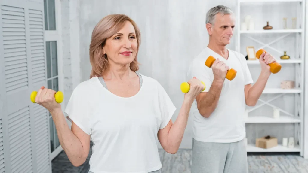 A woman and man in their early 60s performing dumbbell curls together at home, both focused and composed, reflecting the sustainable combined approach to fitness after 50.