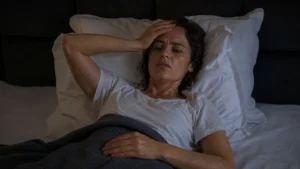 Woman over 50 lying awake in bed at night sweating with hand on forehead, experiencing night sweats and sleep disruption after 50