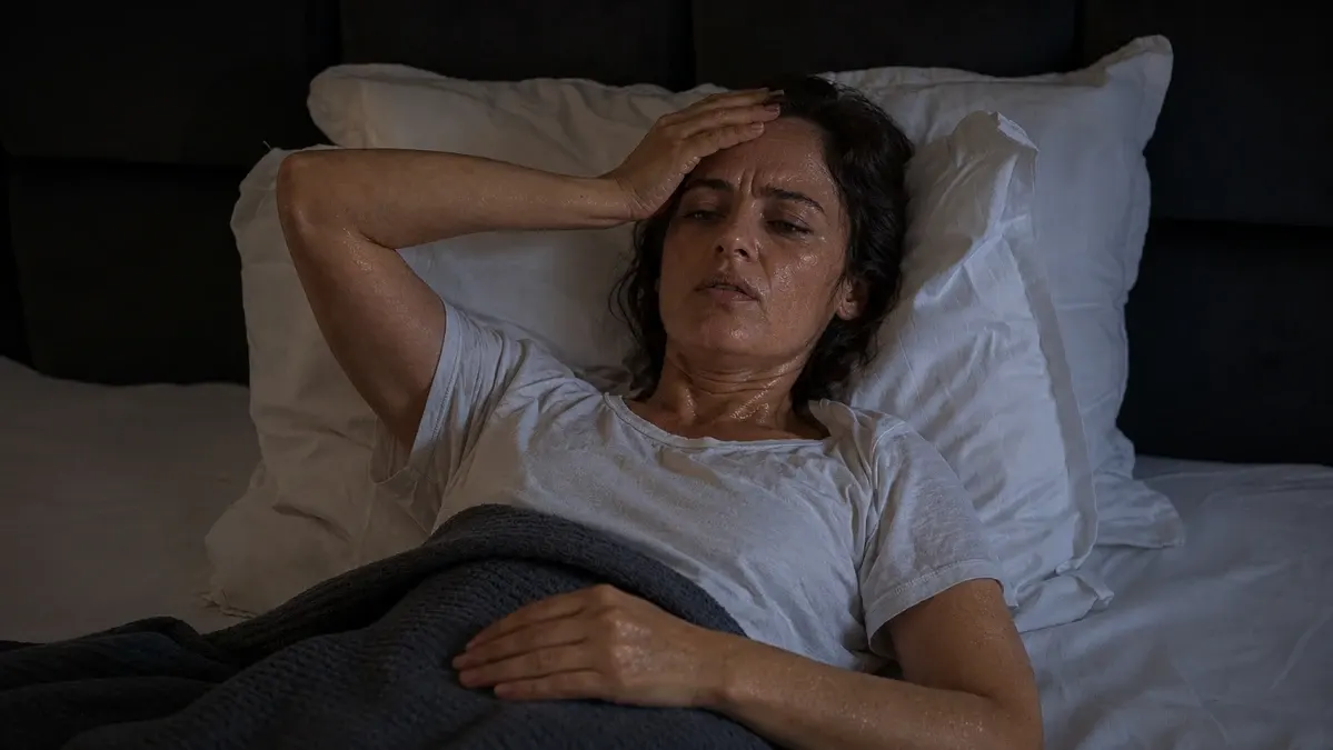 Woman over 50 lying awake in bed at night sweating with hand on forehead, experiencing night sweats and sleep disruption after 50
