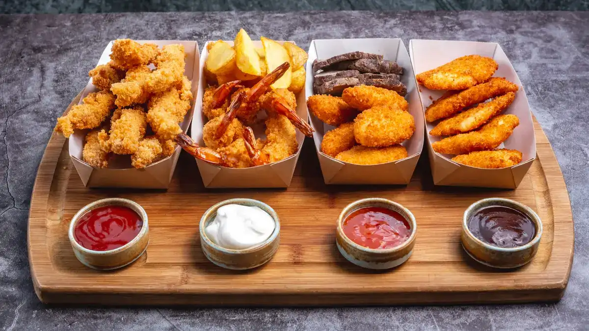 An assortment of fried appetizers including breaded chicken strips, fried shrimp, and potato wedges served in paper containers with four dipping sauces. A visual example of high-trans-fat foods that can trigger inflammation.