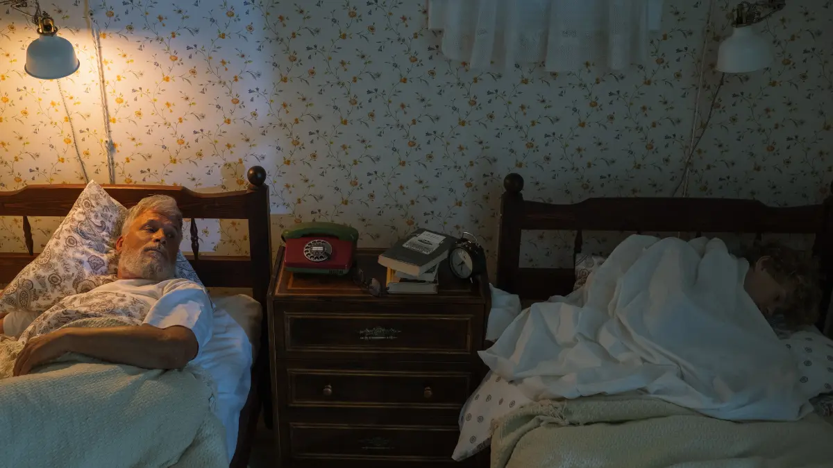 Older man lying awake at night in bedroom while partner sleeps, experiencing common age-related insomnia and sleep disruption after 50.