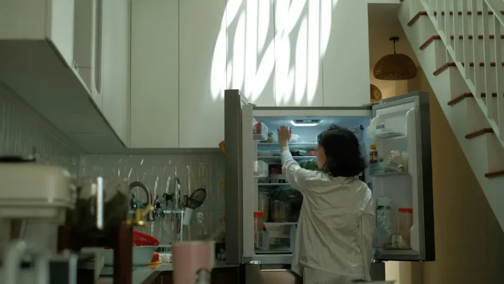 A woman in her late 50s reaches into a refrigerator for a jar of plain kefir in a naturally lit kitchen.