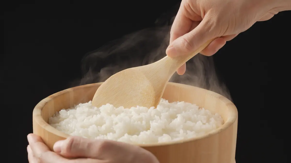 A person holds a warm wooden bowl of freshly cooked, steaming white rice and scoops it with a flat wooden paddle.