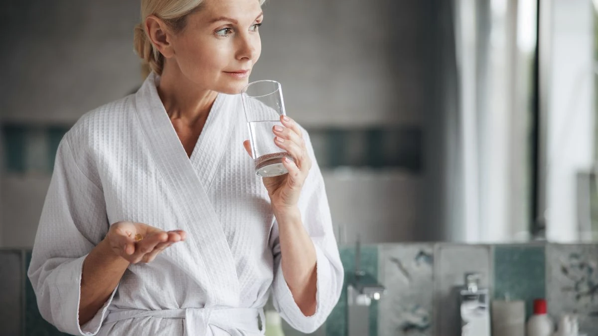 A middle-aged woman in a white bathrobe holding a glass of water and several small vitamin D gel capsules in her palm.