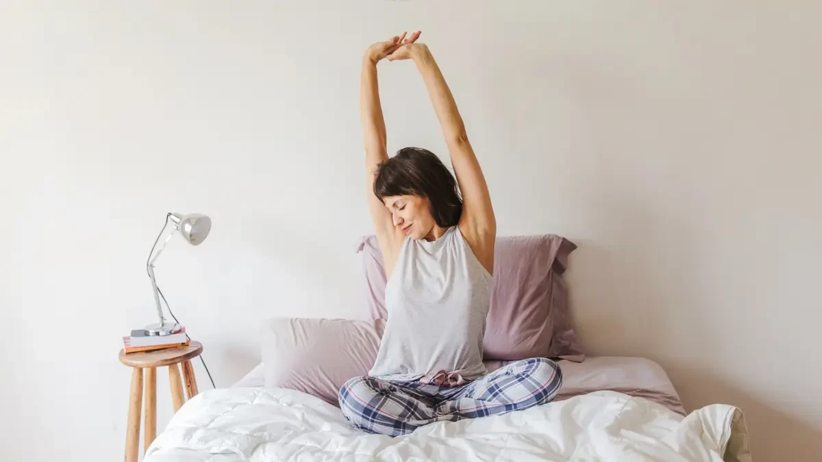 Mature woman sitting on her bed and stretching with a peaceful expression to illustrate the reduction in physical heaviness as mitochondrial density begins to rebuild after 50.
