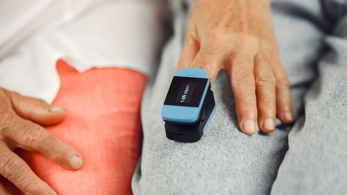 Older adult's hand resting on grey fabric with a blue pulse oximeter clipped to the index finger, monitoring blood oxygen levels at home.
