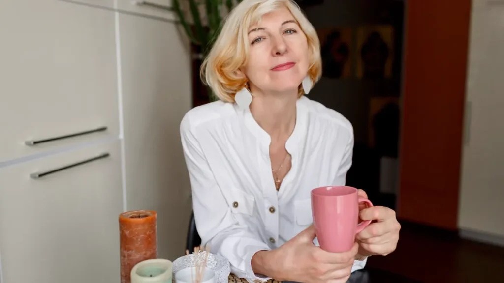 Mature woman with short blonde hair holding a pink coffee mug at a kitchen table, representing hormonal changes and caffeine sensitivity after menopause.