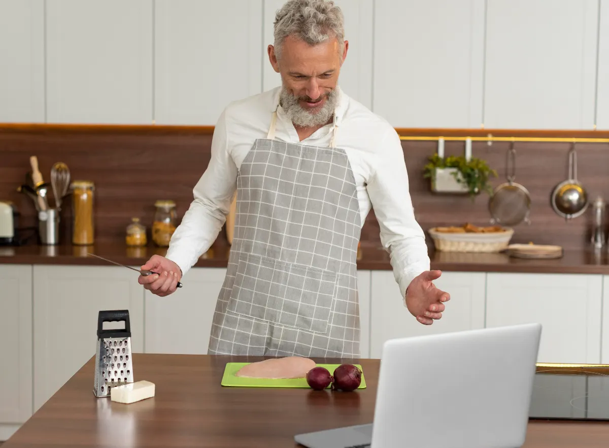 Mature man in a kitchen looking at a laptop with a puzzled expression while trying to follow a simple recipe, representing common brain fog symptoms like poor focus.