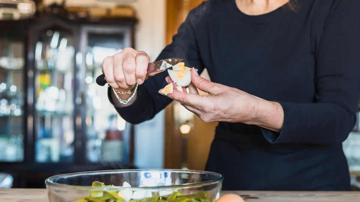 Older woman's hands slicing a hard-boiled egg into a glass bowl of dark green beans, supporting the folate and B12 dietary steps that help lower homocysteine.