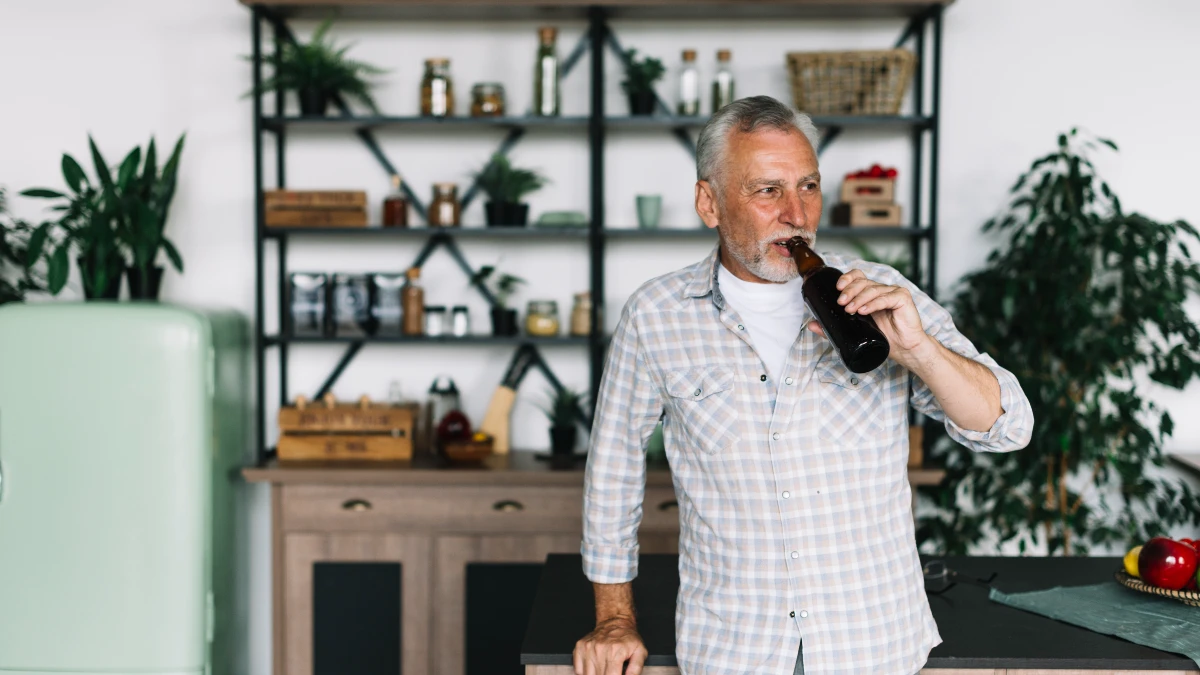 Older man drinking a beer bottle in a home kitchen — regular alcohol consumption forces the kidneys to deprioritize uric acid clearance, allowing levels to build.