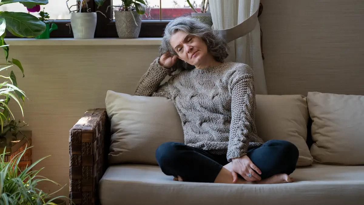 Silver-haired woman in her early 60s sitting cross-legged on a sofa with eyes closed and head resting against her hand, reflecting the unexplained fatigue associated with B12 deficiency.