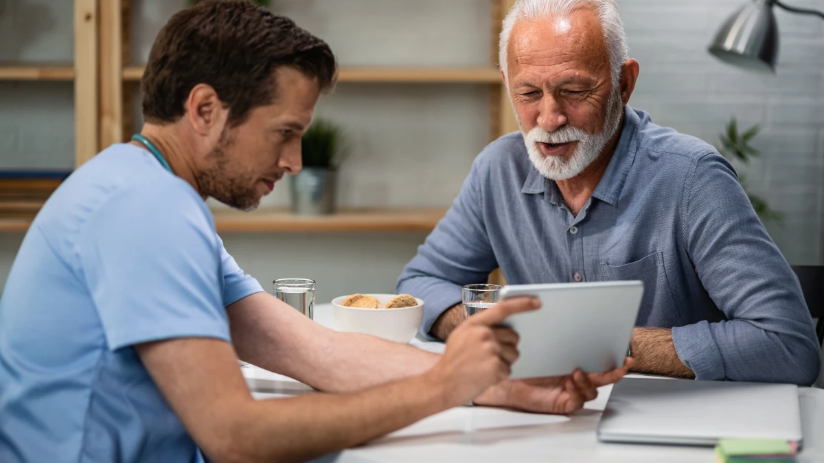 Senior man reviewing medical test results on a tablet with a healthcare practitioner at a home table — tracking uric acid and metabolic numbers over time to catch changes before symptoms appear.