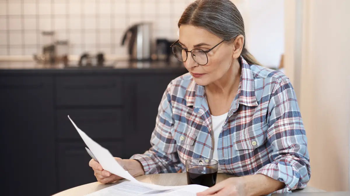 Mature woman in her kitchen reviewing medical paperwork with a pen to prepare for her doctor's appointment regarding B12 status and brain fog symptoms.