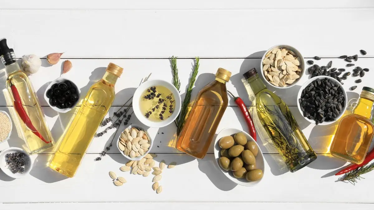Various cooking oil bottles with olives, seeds, and herbs arranged in a flat lay on a white wooden background