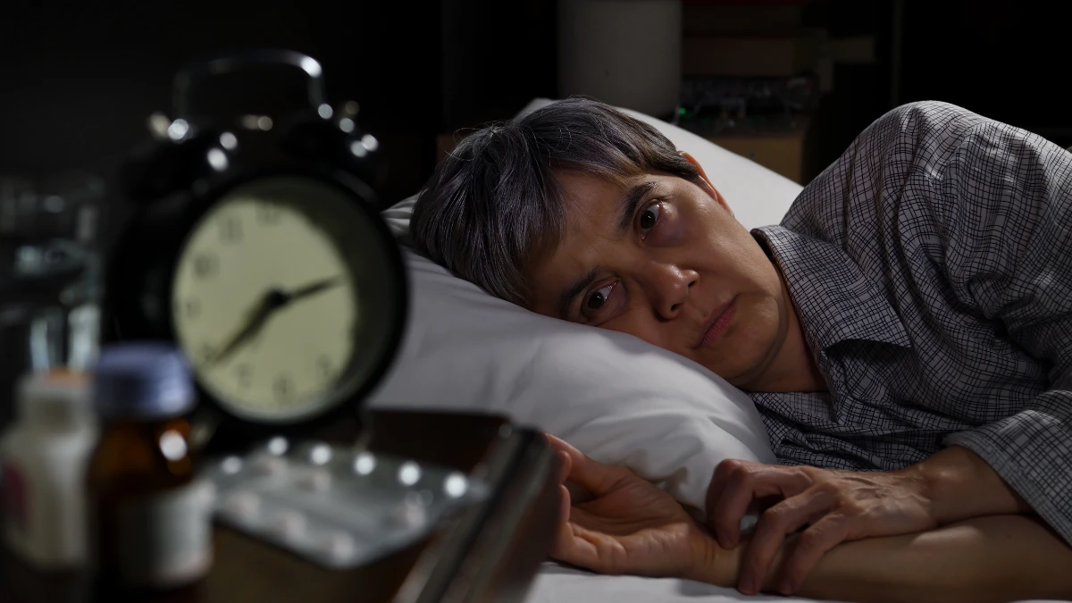 Asian man in his late 50s lying awake in bed at night, staring toward a dim alarm clock on the nightstand beside supplement bottles, unable to sleep as caffeine consumed hours earlier remains active in his system after 50.