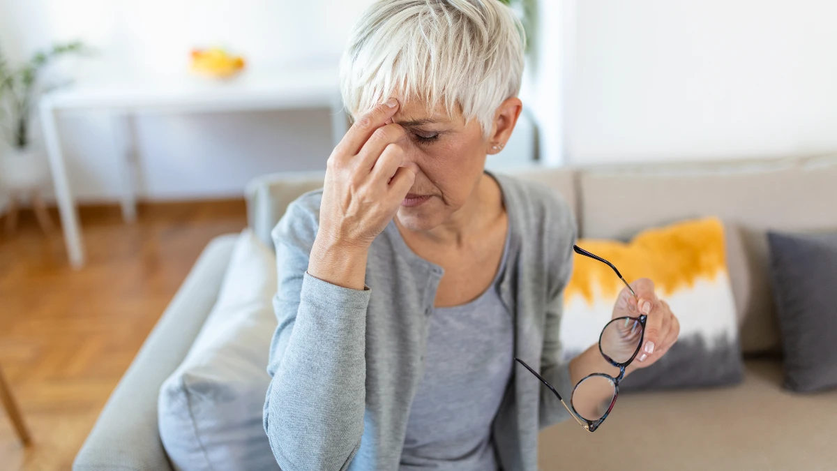 Senior woman in her 60s removing her glasses and rubbing her eyes while sitting on a sofa, showing discomfort from dry eye symptoms common after age 50.