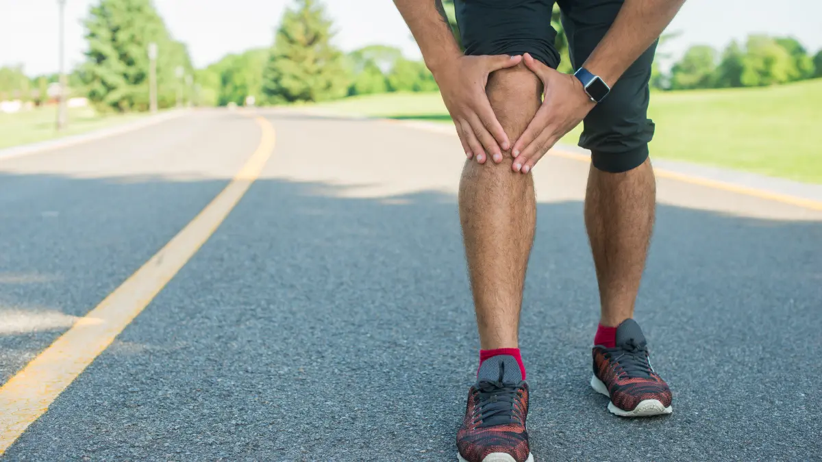 Mature man in activewear standing on a paved walking path while clutching his sore knee in pain.