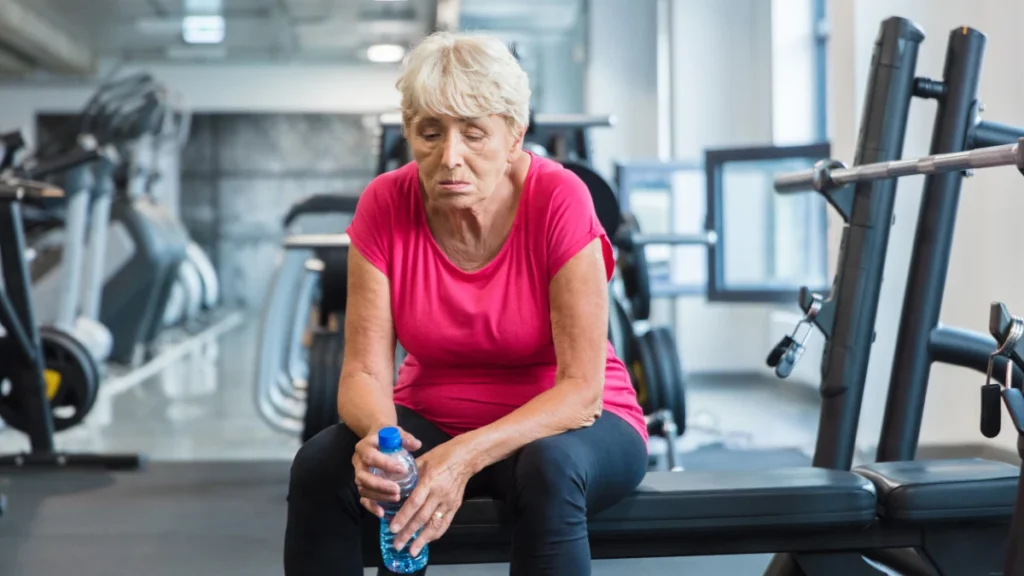 Exhausted senior woman resting on gym bench after workout showing why many adults over 50 struggle with gym consistency and burnout.