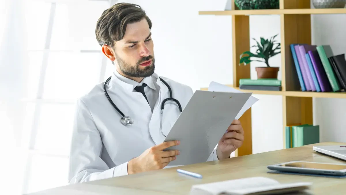 Male doctor in white coat carefully reviewing medical documents at his office desk with bookshelves behind him.