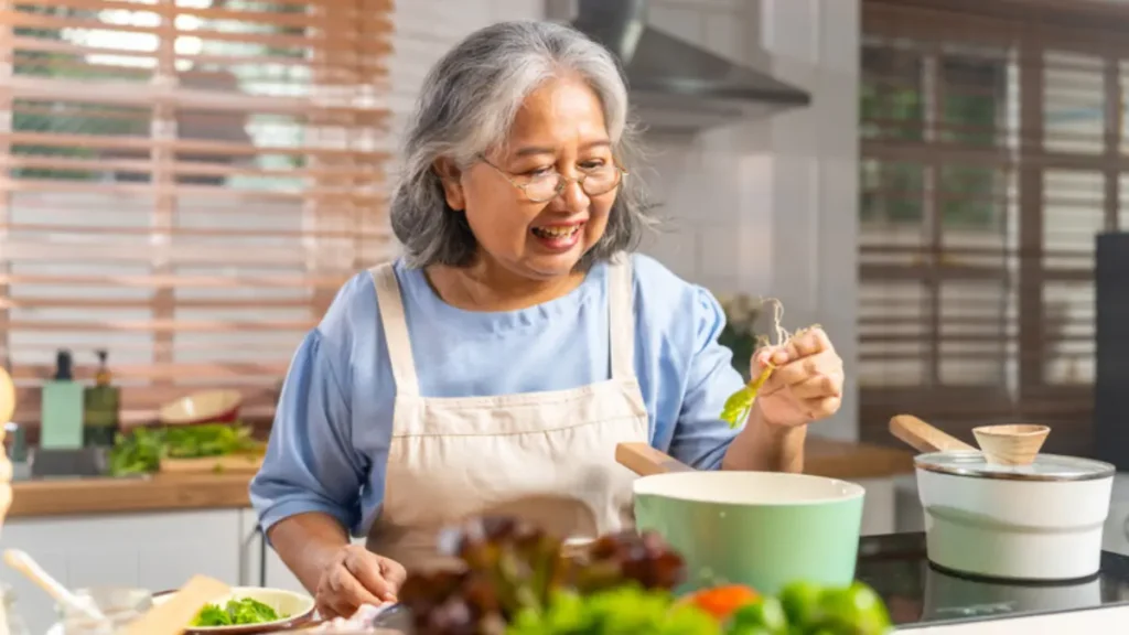 Asian woman in her 60s adding fresh vegetables to a pot while preparing a nutrient-dense meal at home, pairing whole foods with bone broth to improve gut absorption after 50.