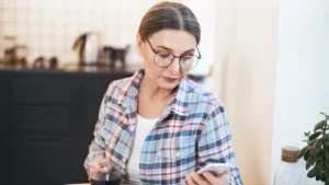 Silver-haired woman in her late 50s sitting at a kitchen table with two glass mugs of tea, smiling softly at her phone while reviewing her homocysteine test results.