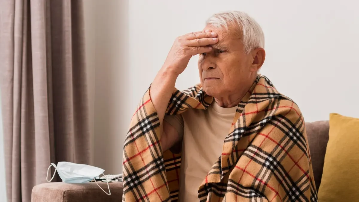 Older man wrapped in blanket on sofa illustrating post-illness fatigue and muscle weakness from anabolic resistance after 50.