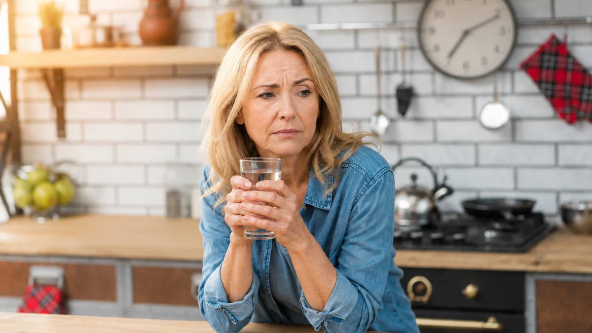 Woman in her 50s holding a glass of water in her kitchen, looking at the wall clock with a frustrated expression during intermittent fasting after 50.