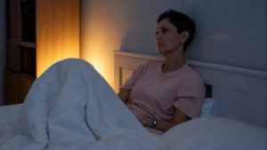 Woman in her 50s sitting awake in a dimly lit bedroom with a sleep tracker on her wrist, staring into the distance with quiet resignation.