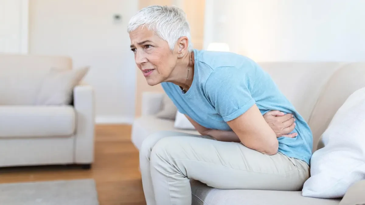 Woman in her 60s sitting on a sofa leaning forward with both hands pressed to her abdomen, experiencing stomach discomfort that may relate to reduced digestive acid after menopause.