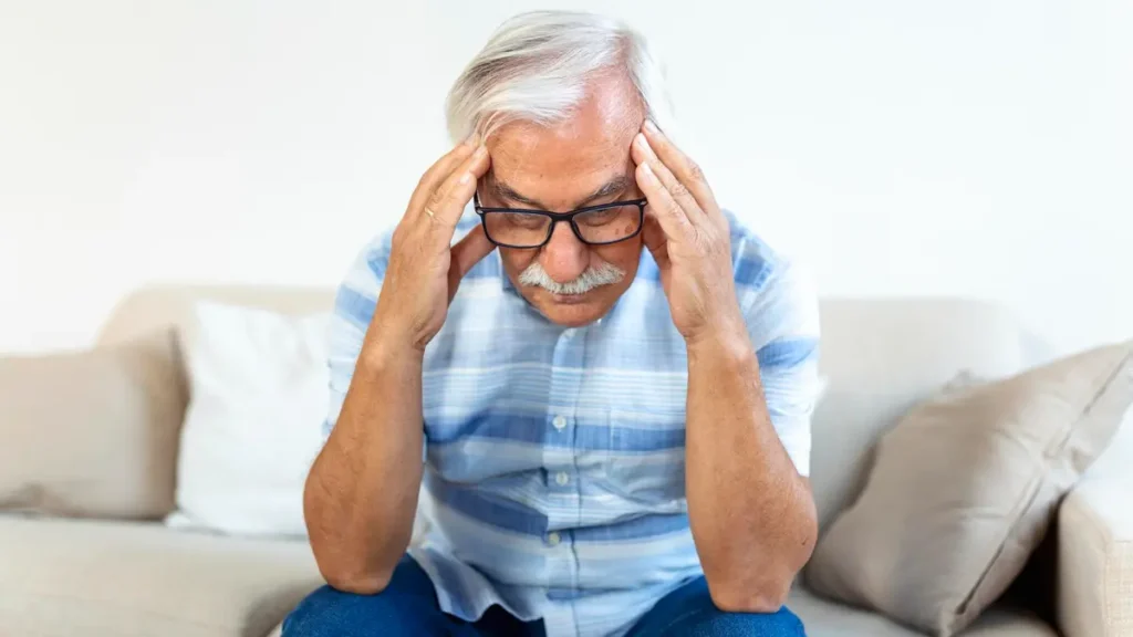 Mature man rubbing his temples on a couch to alleviate morning brain fog caused by age-related protein accumulation inside cells.