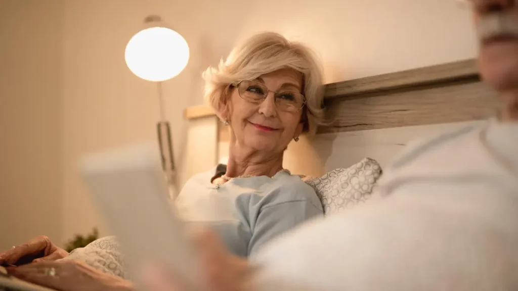 Older woman relaxing in bed at night with a warm lamp, representing a healthy evening routine that protects the overnight fasting window.