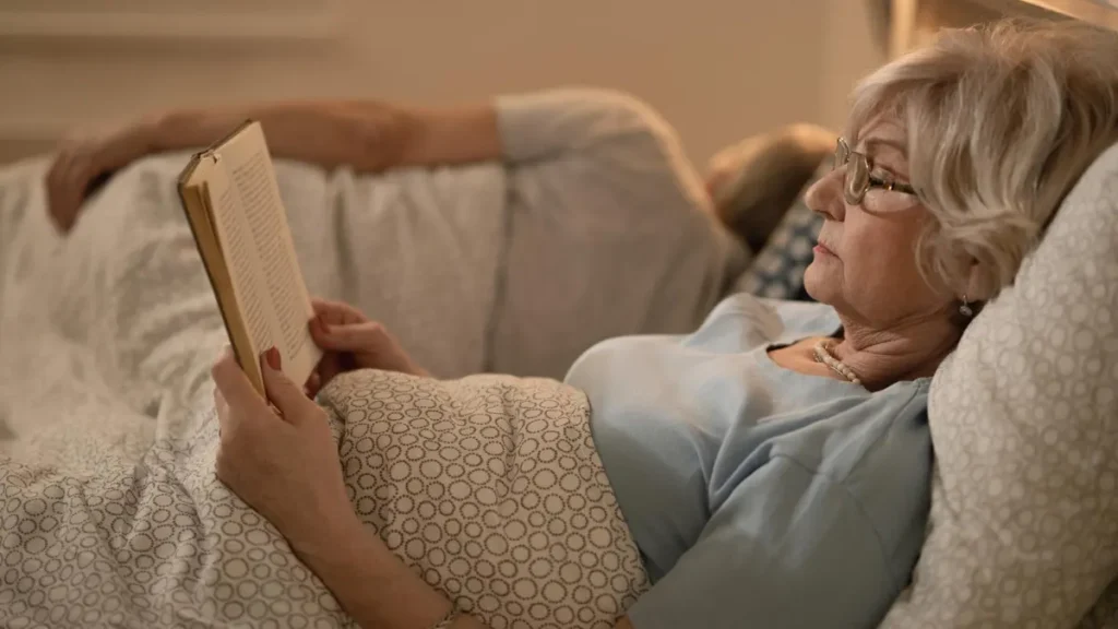 Older woman reading a book in bed under warm lamp light, a screen-free evening routine that helps support better sleep at night.