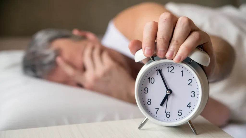 Older man lying in bed reaching to turn off white alarm clock after waking up tired despite getting 8 hours of sleep after 55.