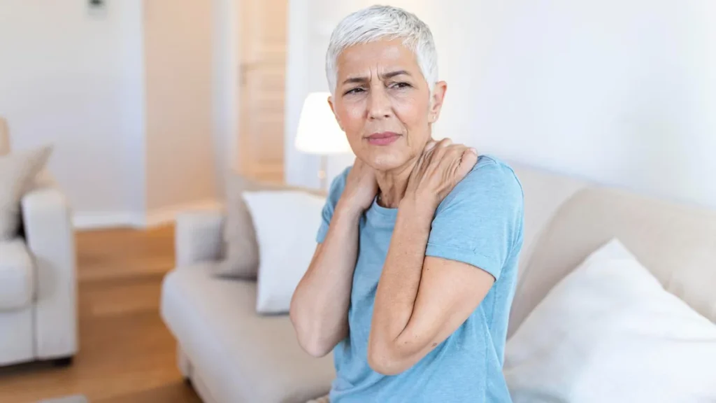 An older woman holding her sore shoulder at home, reflecting the lingering muscle soreness that persists for days after exercise in adults over 50.