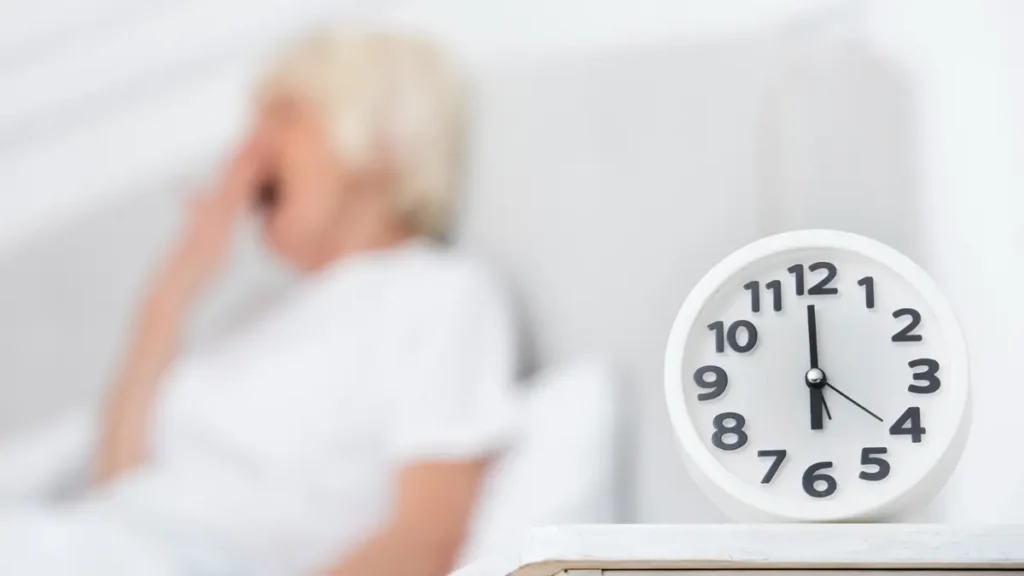 Yawning older woman sitting up in bed beside an alarm clock, reflecting daytime fatigue and brain fog after a poor night's sleep.