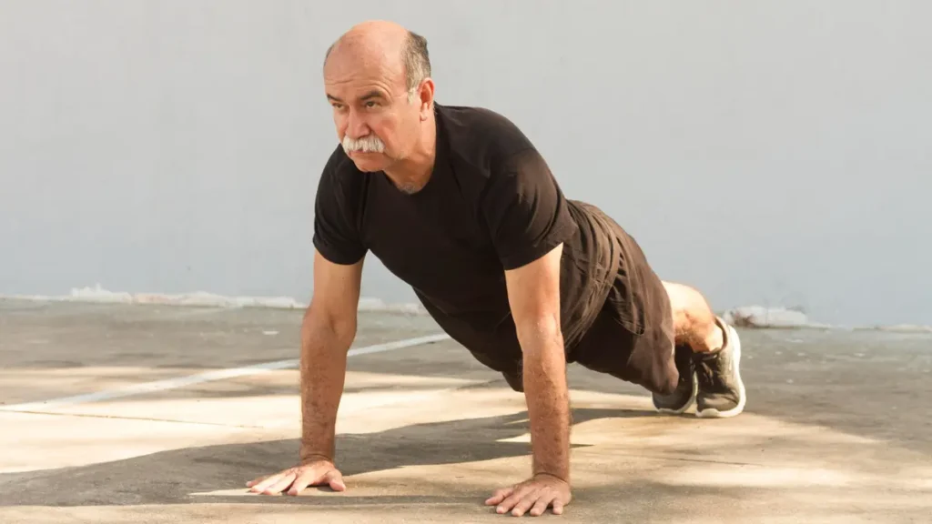 A fit older man in a black t-shirt performing a push-up on concrete pavement, demonstrating resistance training as part of muscle recovery after 50.