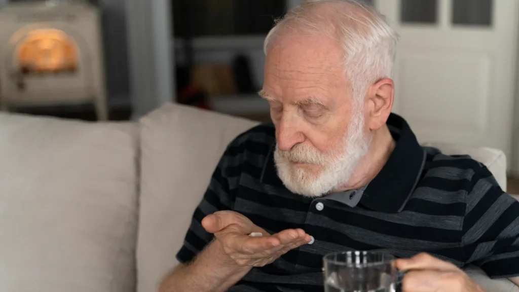 Senior man holding medication pill with water glass at home, showing medication timing as a hidden cause of afternoon fatigue after 50