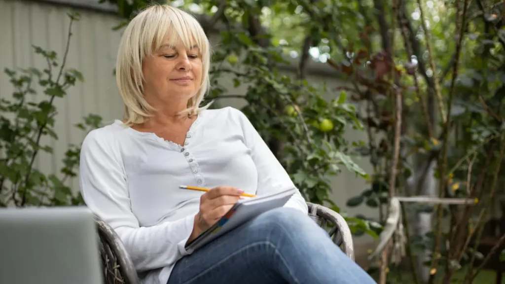 Calm mature woman writing in a journal outdoors in a garden, demonstrating late morning focus and stable energy levels after two weeks.