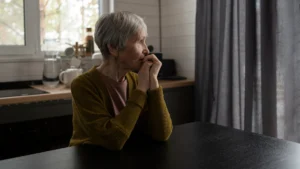 Older woman sits alone at a kitchen table, hands clasped, gazing out a window, reflecting on loneliness and heart health after 50.