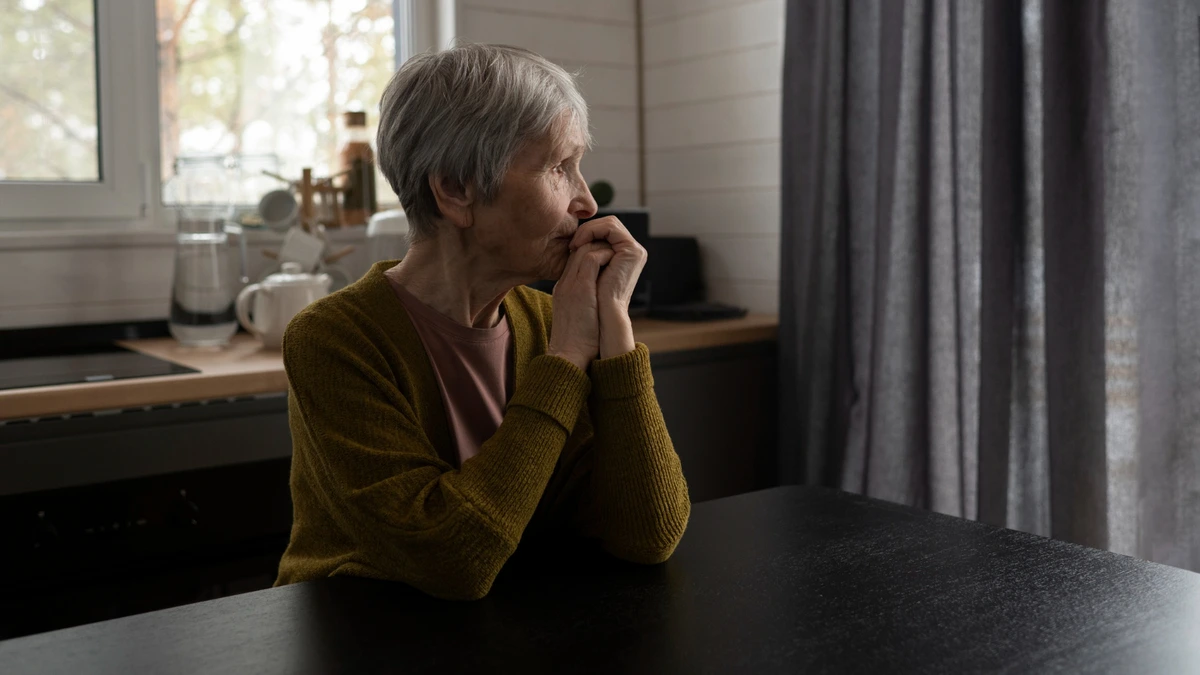 Older woman sits alone at a kitchen table, hands clasped, gazing out a window, reflecting on loneliness and heart health after 50.