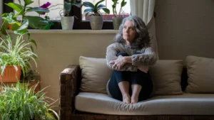 Mature woman with silver hair sitting on a couch hugging her knees, looking upward with a thoughtful, analytical expression to mentally retrace her steps during a moment of memory lapses after 50.