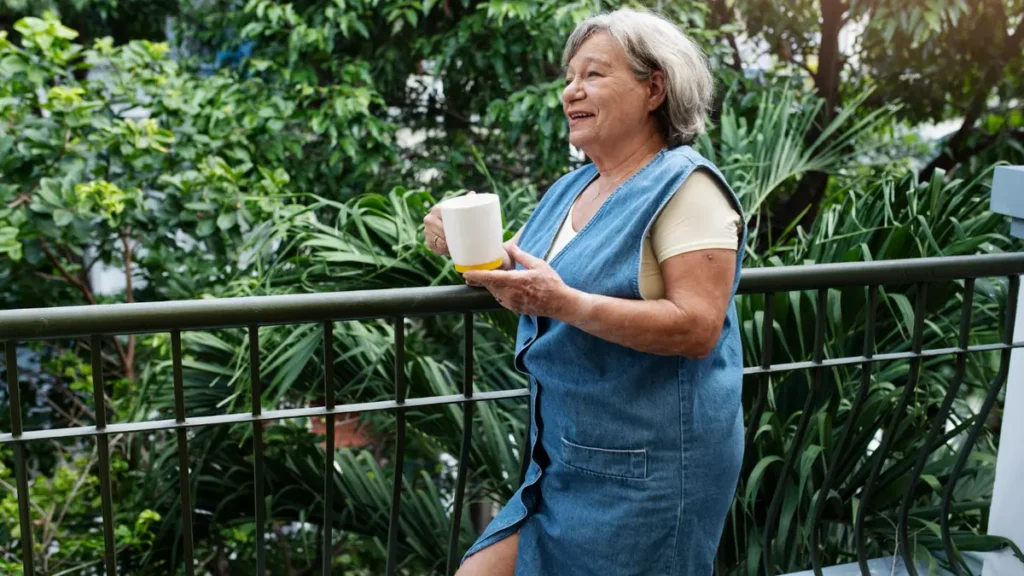 Mature woman standing on a lush green balcony holding a coffee mug to get natural morning light and stabilize her circadian rhythm.