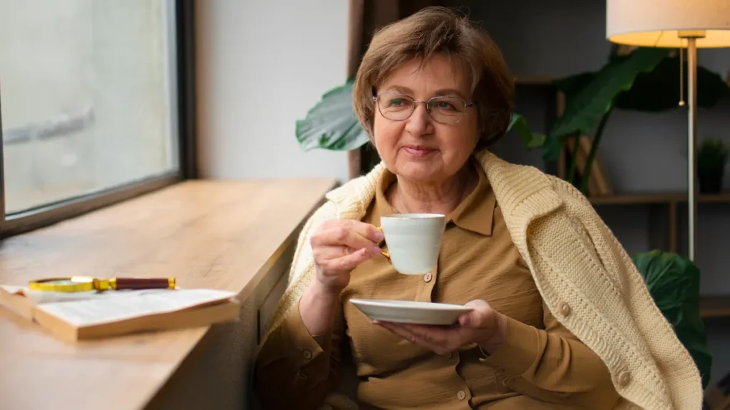 Woman in her late 60s sitting quietly by a window holding a cup of tea with a book nearby, illustrating the five-minute pre-meal stress reset that lowers cortisol and improves insulin sensitivity before eating.