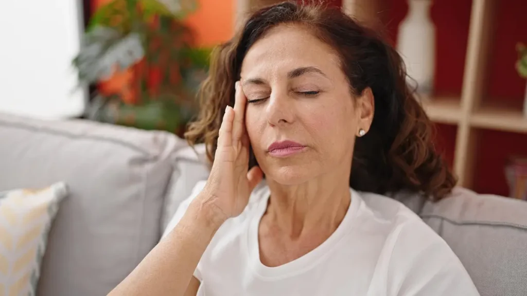 Mature woman sitting on a living room sofa pressing her hand to her temple to manage a persistent headache linked to systemic polymyalgia rheumatica symptoms.