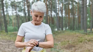 Exercise and memory after 50 comes into focus as a woman in her sixties checks her fitness tracker mid-walk on a wooded forest trail, pausing to time her routine with a quiet smile.