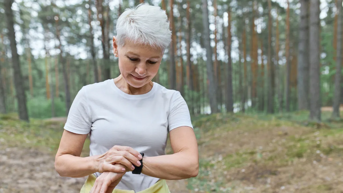 Exercise and memory after 50 comes into focus as a woman in her sixties checks her fitness tracker mid-walk on a wooded forest trail, pausing to time her routine with a quiet smile.