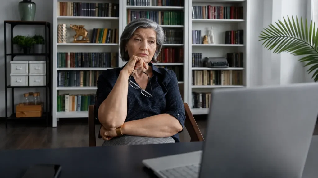 Woman in her late 50s sitting at a home desk in afternoon light with an unfocused expression, reflecting the blood sugar and willpower after 50 afternoon energy crash.