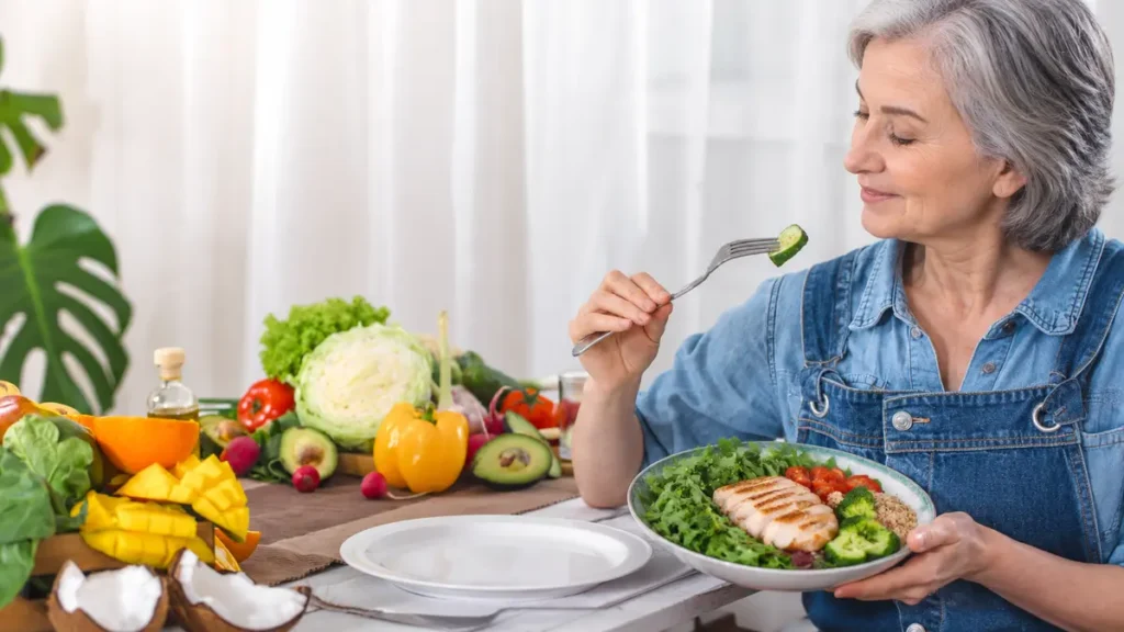 Woman in her late 50s eating a lunch bowl with grilled chicken and leafy greens at a table surrounded by fresh vegetables, reflecting the protein-and-fiber-first meal structure that slows gastric emptying and reduces afternoon blood sugar spikes.